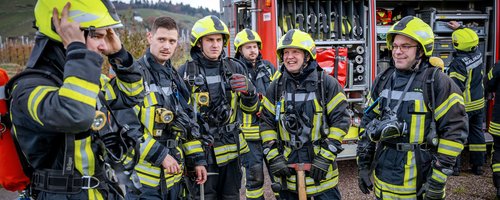 Eine Gruppe von Feuerwehrmännern steht zusammengefasst vor einem Feuerwehrauto. Sie tragen Schutzkleidung und sind mit Ausrüstungen ausgestattet, die sie für ihre Aufgaben als Rettungsleute befähigen.