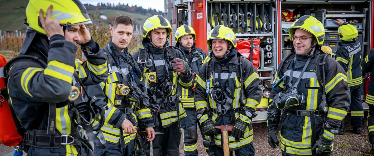 Eine Gruppe von Feuerwehrmännern steht zusammengefasst vor einem Feuerwehrauto. Sie tragen Schutzkleidung und sind mit Ausrüstungen ausgestattet, die sie für ihre Aufgaben als Rettungsleute befähigen.