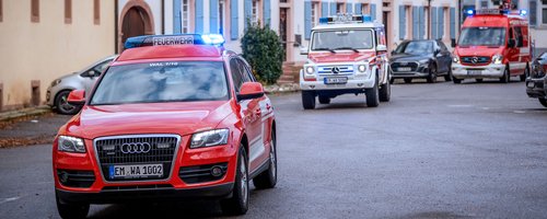 Feuerwehrfahrzeuge unterwegs in einer Stadtstraße, mit Blaulicht und Einsatzfahrzeugen im Vordergrund
