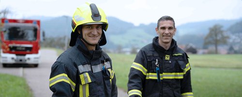 Zwei Feuerwehrleute in Einsatzkleidung stehen auf einem Weg, während ein Feuerwehrfahrzeug im Hintergrund sichtbar ist.