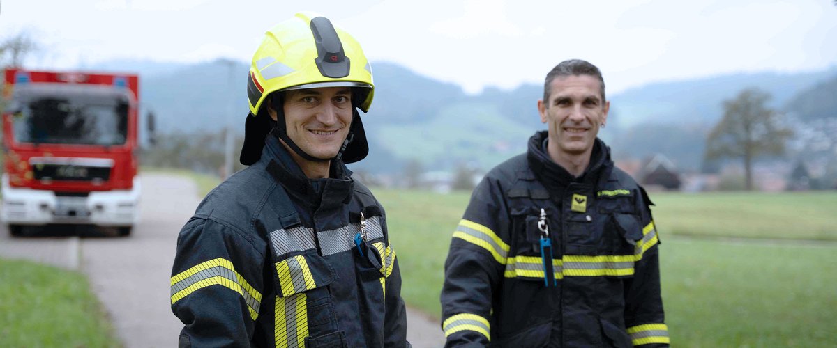 Zwei Feuerwehrleute in Einsatzkleidung stehen auf einem Weg, während ein Feuerwehrfahrzeug im Hintergrund sichtbar ist.