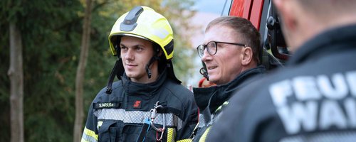 Zwei Feuerwehrleute in Schutzkleidung stehen neben einem Einsatzfahrzeug im Freien.