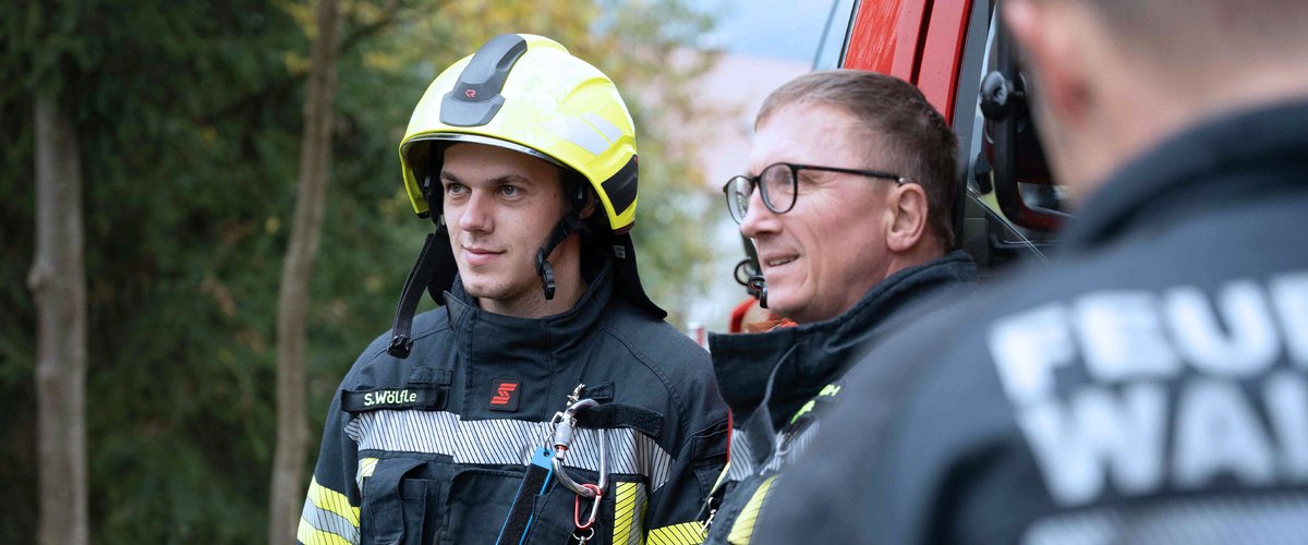 Zwei Feuerwehrleute in Schutzkleidung stehen neben einem Einsatzfahrzeug im Freien.