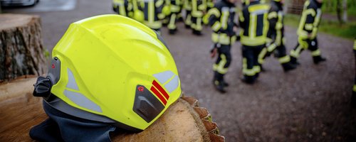 Gelber Feuerwehrhelm auf einem Baumstumpf, im Hintergrund Feuerwehrleute in Einsatzkleidung