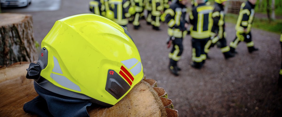 Gelber Feuerwehrhelm auf einem Baumstumpf, im Hintergrund Feuerwehrleute in Einsatzkleidung