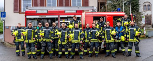 Feuerwehrübung vor einem zweigeschossigen Haus mit Feuerwehrfahrzeug und Einsatzkräften in Schutzkleidung, während ein Feuerwehrmann über eine Leiter in ein Fenster steigt. Im Hintergrund ist ein Kirchturm sichtbar.