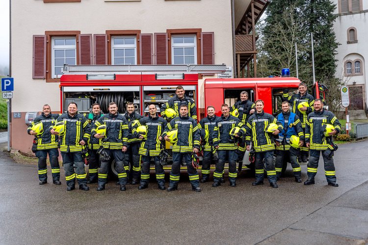 Feuerwehrübung vor einem zweigeschossigen Haus mit Feuerwehrfahrzeug und Einsatzkräften in Schutzkleidung, während ein Feuerwehrmann über eine Leiter in ein Fenster steigt. Im Hintergrund ist ein Kirchturm sichtbar.