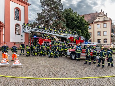 Gruppenfoto der Feuerwehr mit Löschfahrzeug und Einsatzkräften in Uniform vor historischer Kulisse